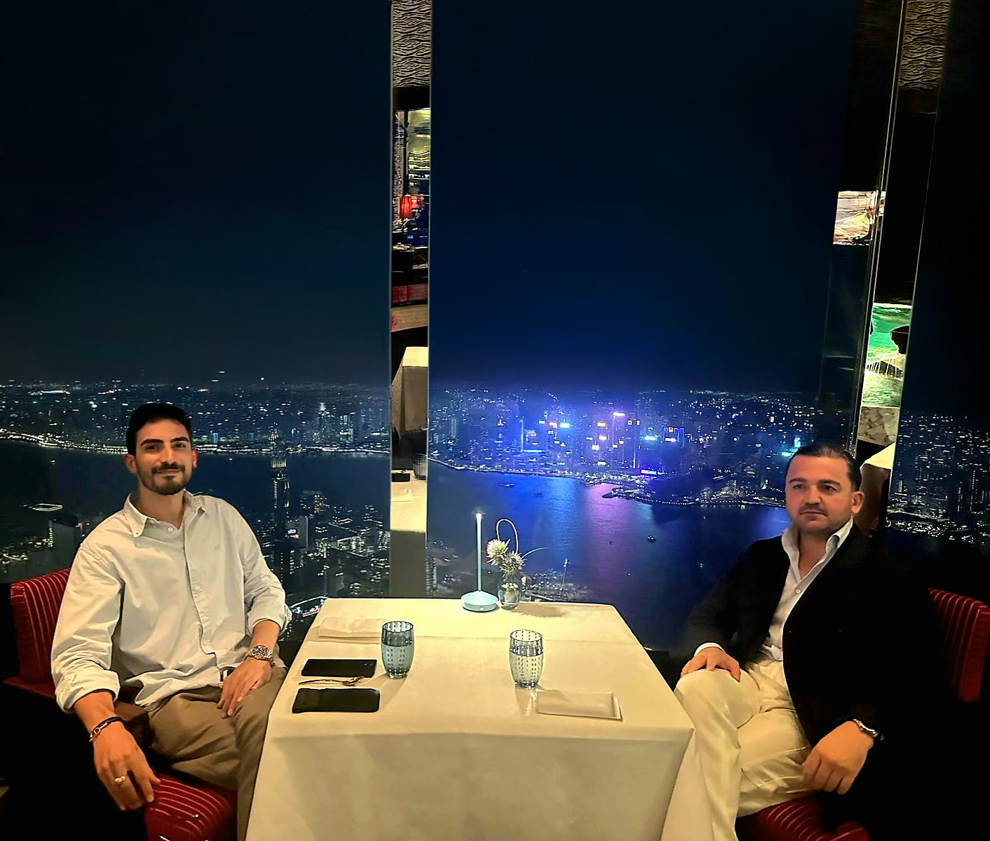 The two partners of Ariana Precious Stones at a restaurant table, a harbour skyline at night behind them